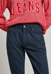 Czerwona bluza z napisem "JEANS" w białym kolorze, zestawiona z ciemnogranatowymi spodniami z przodu z kieszeniami i zapięciem na guzik. Gładka struktura materiału.
