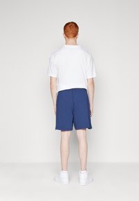 Maglietta bianca a girocollo con maniche corte, abbinata a pantaloni corti di cotone blu e sneaker alte bianche; vista dal retro.