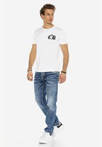Weißes T-Shirt mit kurzen Ärmeln und schwarzem Logo auf der linken Brust, kombiniert mit hellblauen Jeans und weißen Sneakern mit schwarzen Akzenten.