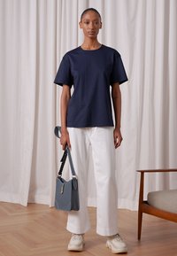 Camiseta de manga curta azul-marinho combinada com calças largas brancas, segurando uma bolsa azul com alça trançada e usando ténis brancos.