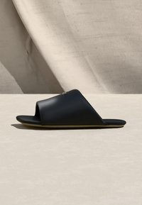 Μαύρο ανοιχτό σανδάλι τύπου slide με φαρδιά λεία λουράκι και επίπεδη σόλα, εκτεθειμένο σε μπεζ επιφάνεια με ουδέτερο υφασμάτινο φόντο.