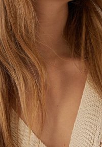 Nahaufnahme eines Halses und oberen Brustbereichs einer Person mit langen, gewellten, hellbraunen Haaren und einem beigefarbenen Strickoberteil.