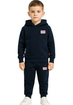 Jeune garçon debout de face portant un sweat à capuche bleu marine et un pantalon de survêtement avec des baskets blanches sur fond blanc.