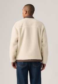 Chaqueta de forro polar color crema con un exterior texturizado, cuello marrón y un corte relajado, combinada con jeans de mezclilla azules oscuros.