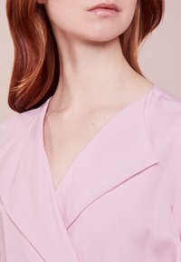 Roodharig persoon in een lichtroze V-hals top, die de nek, kin en een deel van de lippen toont tegen een neutrale achtergrond.