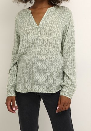 Femme portant un chemisier à manches longues vert clair avec un motif de feuilles blanches et un jean noir, debout devant un fond beige uni.