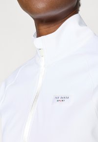 Balta sportinė užtrauktuku užsegama striukė su aukštu apykaklės kraštu, turinti stačiakampį prekės ženklo etiketę ir lygų, blizgų paviršių.