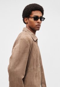 Barna velúr dzseki gallérral, cipzár részlettel. Fekete napszemüveggel viselve. A háttér semleges és letisztult.