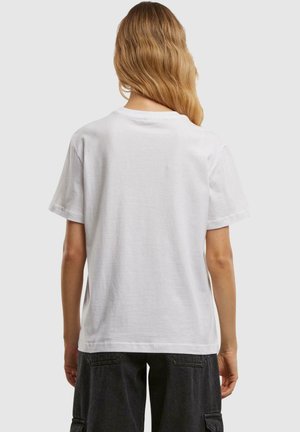 En blond person iført en ensfarvet hvid kortærmet t-shirt og sorte cargo shorts, stående med ryggen mod beskueren.