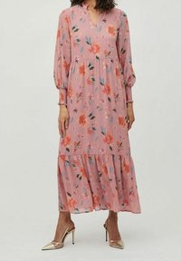 Femme portant une longue robe rose à fleurs avec des manches longues et un ourlet à volants, associée à des chaussures métalliques à talons aiguilles pointues.