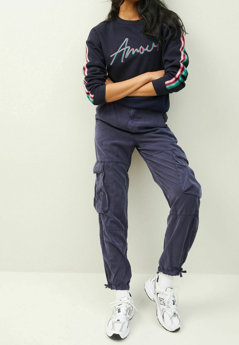 Personne portant un pantalon cargo bleu marine foncé, un t-shirt noir à manches longues avec des bandes colorées sur les manches et des baskets blanches, debout les bras croisés.