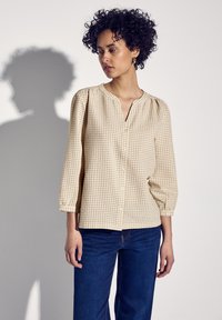 Camicia beige a quadri con maniche a tre quarti e scollo a V, abbinata a jeans a palazzo blu scuro. Tessuto liscio.