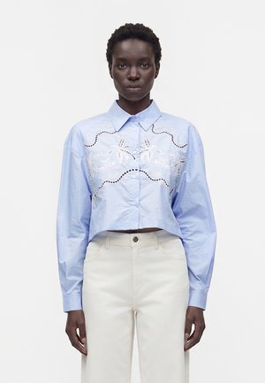 Femme portant une chemise courte bleu clair à boutons avec broderie florale blanche et pantalon taille haute crème, debout devant un fond blanc.
