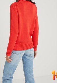 Maglione rosso lavorato a maglia con colletto alto e orlo a coste. Abbinato a jeans azzurri dal taglio rilassato. Nessun motivo visibile o dettagli in metallo.