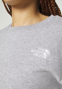 Gros plan d'une personne portant un pull gris clair à col rond avec le logo blanc « The North Face » brodé sur la poitrine.