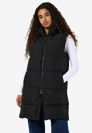 Femme aux longs cheveux bouclés portant un gilet sans manches rembourré noir par-dessus un haut blanc à manches longues et un jean bleu, les mains dans les poches du gilet.