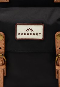 Black fabric backpack featuring a white label with "DOUGHNUT" in brown text, brown leather straps, and brass hardware accents.