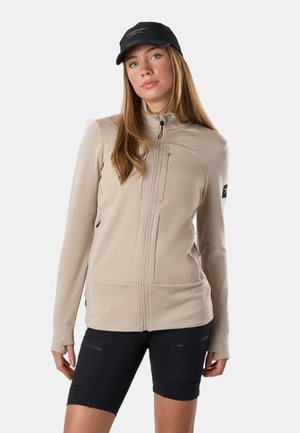 Frau mit langen Haaren, die eine beige Jacke mit Reißverschluss und schwarze Shorts trägt, steht vor einem schlichten Hintergrund, mit schwarzer Mütze auf dem Kopf.