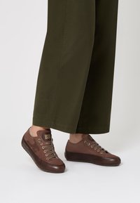 Scarpe casual marroni con lacci e suole scure indossate con pantaloni larghi verde oliva, su uno sfondo chiaro e neutro.