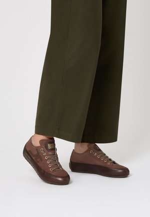 Scarpe casual marroni con lacci e suole scure indossate con pantaloni larghi verde oliva, su uno sfondo chiaro e neutro.