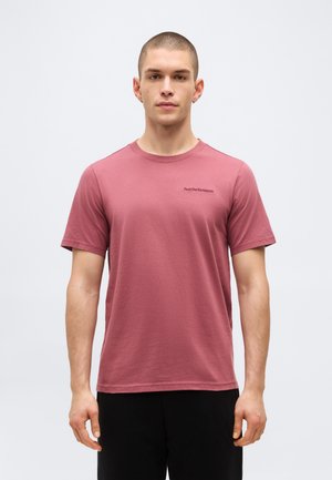 Moški s kratkimi lasmi, oblečen v rožnato kratko majico z okroglim izrezom in črne hlače, stoji pred enobarvnim ozadjem.