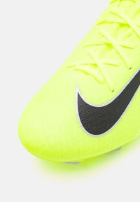 Kirkkaankeltainen jalkapallon kengät, joissa on teksturoitu verkko-yläosa, musta Nike-logo ja hopeiset yksityiskohdat. Narut sijaitsevat keskeisesti.