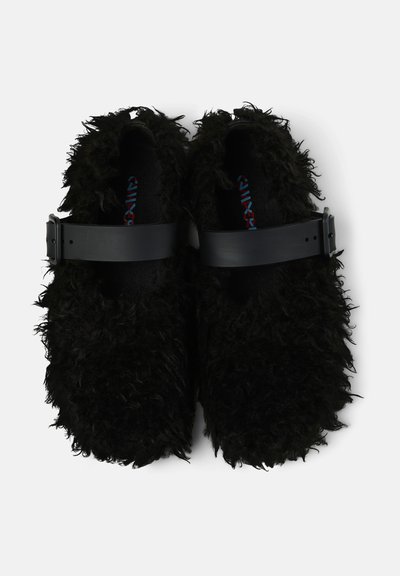 Scarpe nere in peluche senza lacci con una larga fascia nera regolabile sulla parte superiore, viste dall'alto su uno sfondo bianco.