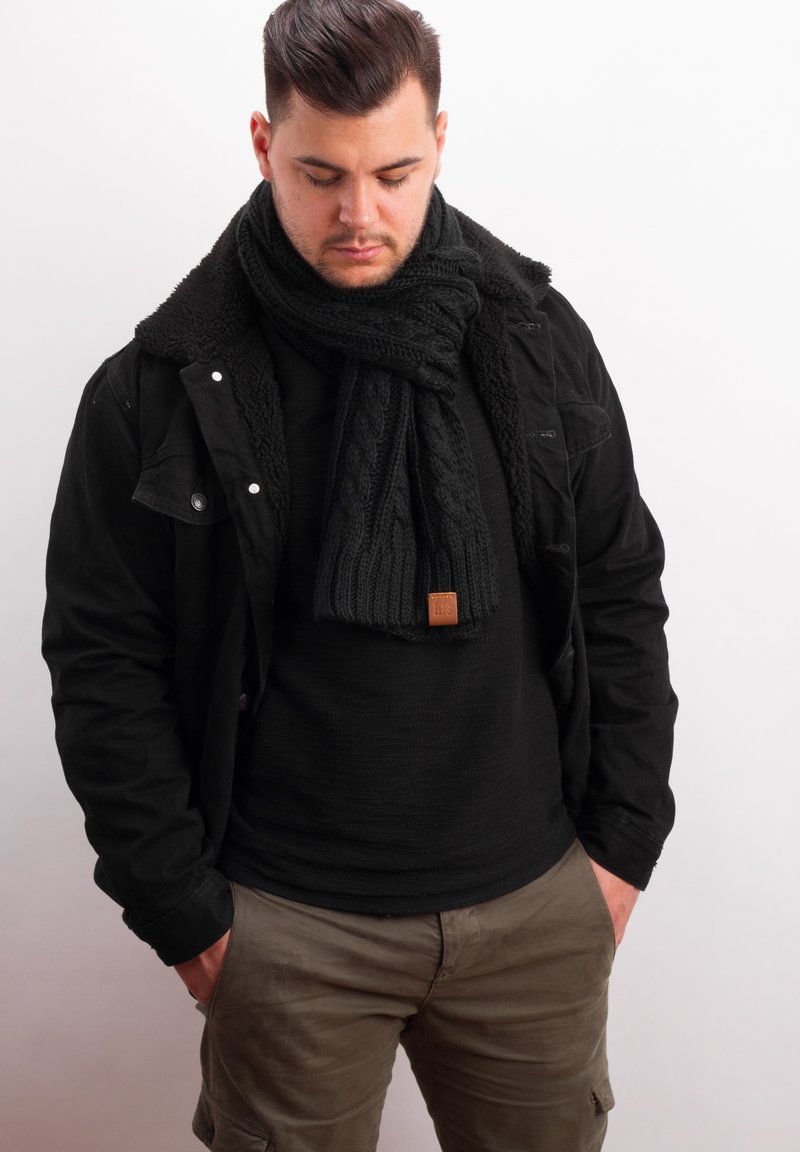 Black knitted scarf with a textured pattern, worn over a black sweater, layered with a black jacket featuring a sherpa collar.