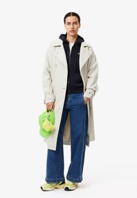 Light beige trench coat over navy hoodie, wide blue jeans, and neon green sneakers. Holding a green cap and a small mesh handbag.