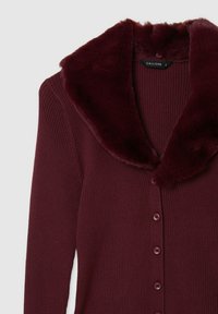 Cardigan borgogna a coste con colletto in pelliccia finta, dotato di bottoni frontali e un design aderente. Texture morbida e aspetto caldo.