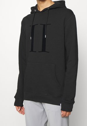 Persona que lleva una sudadera con capucha negra con una gran letra oscura "H" en el pecho y pantalones gris claro sobre un fondo blanco liso.