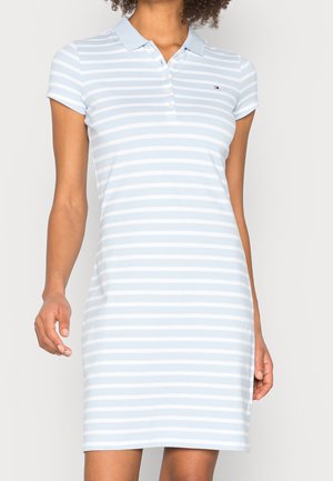 Kobieta ubrana w jasnoniebieską i białą, poziomo prążkowaną, krótkim rękawem sukienkę polo z małym logo na piersi.
