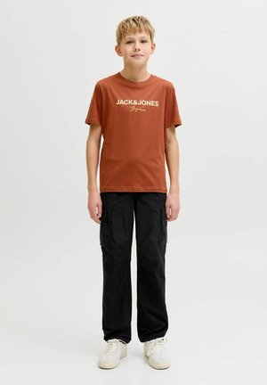 Dreng stående iført rustfarvet Jack & Jones t-shirt, sorte cargo bukser og hvide sneakers mod en ensfarvet lys baggrund.