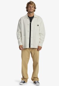 White button-up jacket with a chest pocket, worn over a black shirt; paired with beige pants and black sneakers.