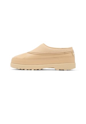 Beige slip-on sko med tyk sål og tekstureret mønster, lav hæl og enkelt syningsdetalje, vist i sideprofil på hvid baggrund.