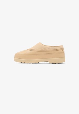 Beige slip-on sko med tyk sål og tekstureret mønster, lav hæl og enkelt syningsdetalje, vist i sideprofil på hvid baggrund.
