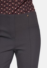 Grau strukturierte Hose mit schmaler Passform und einem kleinen metallischen Logoakzent. Kombiniert mit einem mehrfarbigen, gemusterten Oberteil.