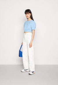 Camisa azul clara de mangas bufantes curto, calças largas brancas, ténis brancos com detalhes em laranja e uma pequena mala azul. Look simples e moderno.