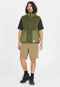 Vihreä fleece-liivi, jossa on korkea kaulus, vetoketjulla suljettava etuosa ja kaksi etutaskua, mustan paidan päällä, yhdistettynä ruskeisiin shortseihin ja mustiin saappaisiin.