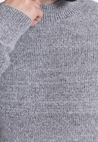 Grauer Strickpullover mit geripptem Muster, hohem Kragen und langen Ärmeln. Der Stoff wirkt weich und leicht fusselig.