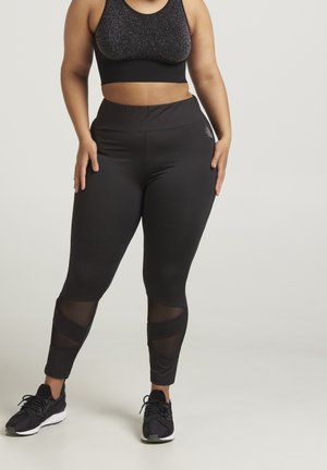 Mujer con leggings negros de cintura alta con paneles de malla y zapatillas negras, de pie contra un fondo claro y liso.