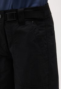 Pantalon noir avec poche latérale et ceinture en sangle noire réglable avec boucle en plastique, porté avec une chemise bleue.