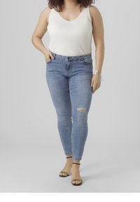 Lehký bílý top bez rukávů s žebrovaným vzorem a hlubokým výstřihem, kombinovaný s modrými slim džíny s roztrhaným kolenem. Černé sandály na tenkých páscích.