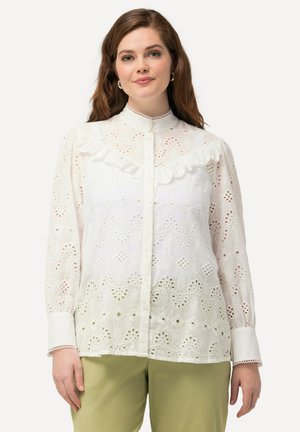 Witte katoenen blouse met ingewikkelde broderie anglaise uitsparingen, een geruffeld kraagje, knoopsluiting en lange mouwen. Gepaarde met olijfgroene broeken.