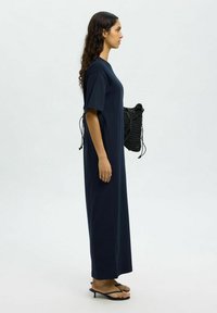 Ilga tamsiai mėlyna suknelė trumpomis rankovėmis, laisvas siluetas, minimalistas dizainas. Modelis laiko juodą pintą maišelį, vilkėdamas juodas šlepetes. Lygus fonas.