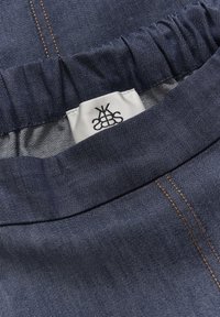 Tăietură din denim bleu închis cu un tiv elastic, având cusături portocalii și o etichetă de îngrijire albă cu text negru vizibil.