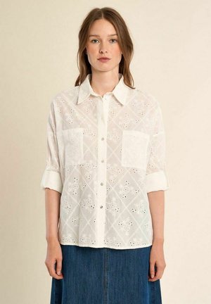 Giovane donna che indossa una camicia bianca in pizzo a occhielli con bottoni e maniche arrotolate e una gonna di jeans blu su sfondo beige.