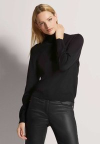 Blonde vrouw in een zwart coltrui en zwarte leren broek, poseert met één hand bij haar hals tegen een egale achtergrond.