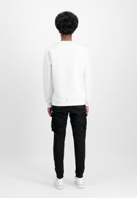 Osoba s kovrčavom kosom obučena u bijelu duksericu, crne cargo hlače i bijele teniske, okrenuta leđima prema jednostavnoj pozadini.