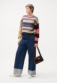 Chunky strikkegenser med flerfargede mønstre, vide blå jeans med grå mansjetter og hvite joggesko. Holder en brun skulderveske.